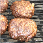 Close-up of three juicy, grilled hamburger patties cooking over charcoal on a grill. The burgers have a rich, browned crust with visible pieces of sautéed onions, and grill marks can be seen underneath.
