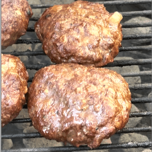 Close-up of three juicy, grilled hamburger patties cooking over charcoal on a grill. The burgers have a rich, browned crust with visible pieces of sautéed onions, and grill marks can be seen underneath.