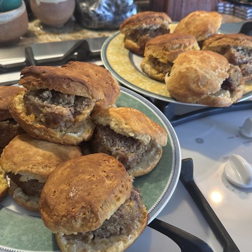 Golden brown sausage biscuits piled on two plates sitting on a stovetop, ready to be served.