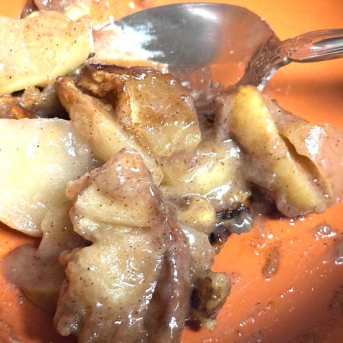 A close-up of warm apple crisp served in a vibrant orange bowl. Sliced, tender apples are coated in a glossy cinnamon glaze and mixed with small clusters of golden crumble topping. A silver spoon is tucked into the side, highlighting the soft, syrupy texture of the dessert.