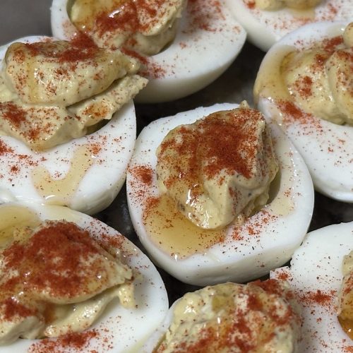 Several deviled egg halves are arranged on a clear plate, each featuring a dollop of textured, creamy yolk filling. The eggs are seasoned with a heavy dusting of bright red paprika, providing a sharp color contrast against the white egg bases. A glossy drizzle of hot honey is visible across each egg, pooling slightly around the filling and adding a sweet, spicy sheen. The close-up shot highlights the varied textures of the smooth egg whites, the soft filling, and the liquid glaze.