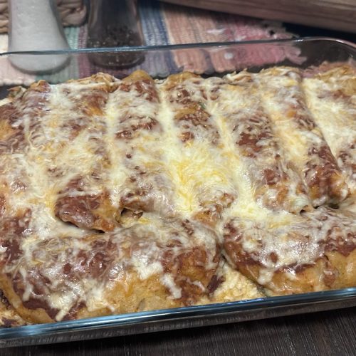 A rectangular glass baking dish is filled with several rolled enchiladas tightly packed together. The enchiladas are coated in a rich red sauce and topped with a thick, melted layer of shredded white and yellow cheese. The dish sits on a dark wood-textured surface, with a salt shaker and pepper grinder visible in the blurred background. Small patches of the golden-brown tortillas and savory filling peek through the bubbly cheese topping.