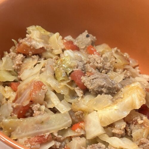 A terracotta orange ceramic bowl is filled with a hearty deconstructed cabbage roll skillet meal. Tender, sautéed green cabbage ribbons are tossed together with crumbled ground meat and diced red tomatoes. Small grains of cooked rice and ground pepper specks are mixed evenly throughout the dish. The hot, savory mixture fills the bowl nearly to the rim in a close-up, angled view.