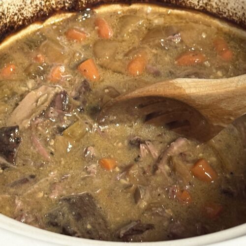 A close-up view shows a hearty pot roast simmering inside a white cooking pot. The dish features tender chunks of braised beef, diced carrots, and soft vegetables submerged in a thick, creamy brown gravy. A wooden slotted spoon rests angled in the center, stirring the mixture. Dark caramelized cooking residue coats the upper interior rim of the pot, highlighting the slow-braising process.
